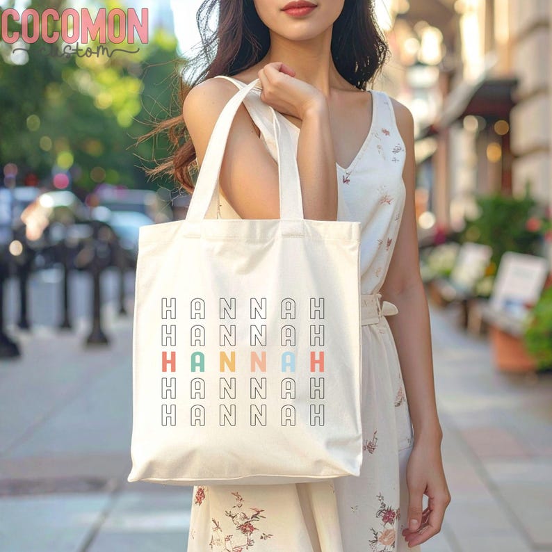 May include: Cream-colored tote bag with the name "HANNAH" printed in a repeating pattern. The name is in a black font, with one instance in red, green, orange, and blue. The bag has a simple design with two handles.