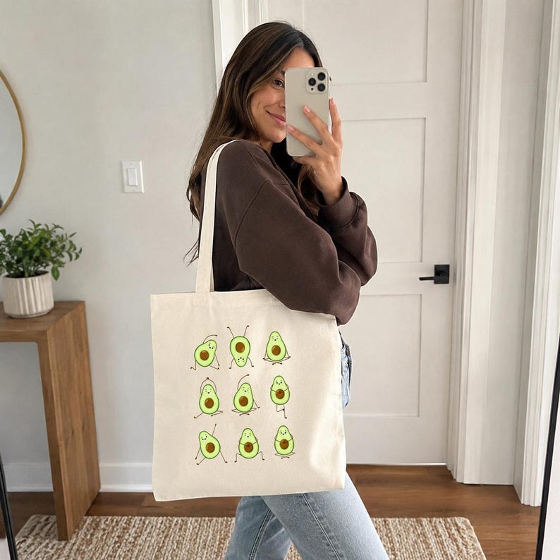 May include: A cream-colored tote bag featuring a repeating pattern of cartoon avocados in various yoga poses. The avocados are green with brown pits and black limbs. The bag has a simple, rectangular shape with long handles.