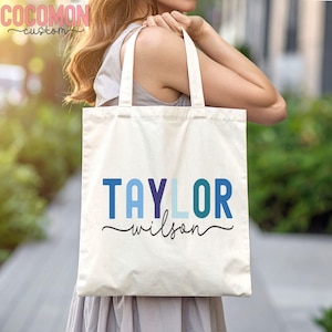 May include: Cream-colored tote bag with the name "TAYLOR WILSON" printed in shades of blue and teal. The word "wilson" is in a cursive font. The bag has two white handles and is made of a sturdy fabric.
