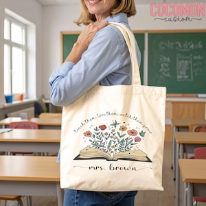 Pu&ograve; includere: Borsa tote color crema con un libro aperto da cui sbocciano fiori colorati. Il testo recita "teach them, love them, watch them grow" e "mrs. Brown". La borsa &egrave; tenuta da una persona in un'aula, con una lavagna sullo sfondo.