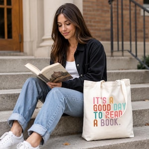 May include: A cream-colored canvas tote bag featuring the multi-colored text "IT'S A GOOD DAY TO READ A BOOK." The bag has two shoulder straps and a rectangular shape, sitting on concrete steps next to a person reading a book.