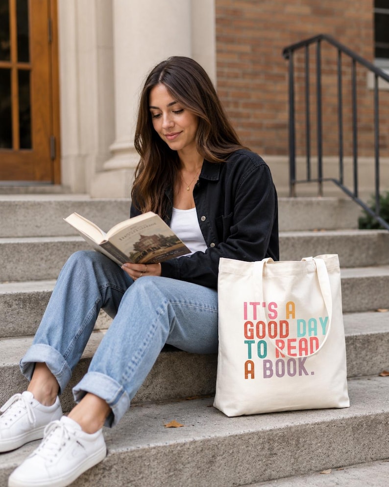 May include: A cream-colored canvas tote bag featuring the multi-colored text "IT'S A GOOD DAY TO READ A BOOK." The bag has two shoulder straps and a rectangular shape, sitting on concrete steps next to a person reading a book.