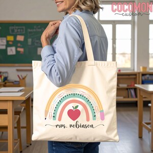 May include: Cream-colored tote bag with a rainbow pencil design and the text "mrs. robinson". The rainbow features a red apple heart, teal, pink, and yellow arcs. The bag has cream-colored handles and is being held by a person.