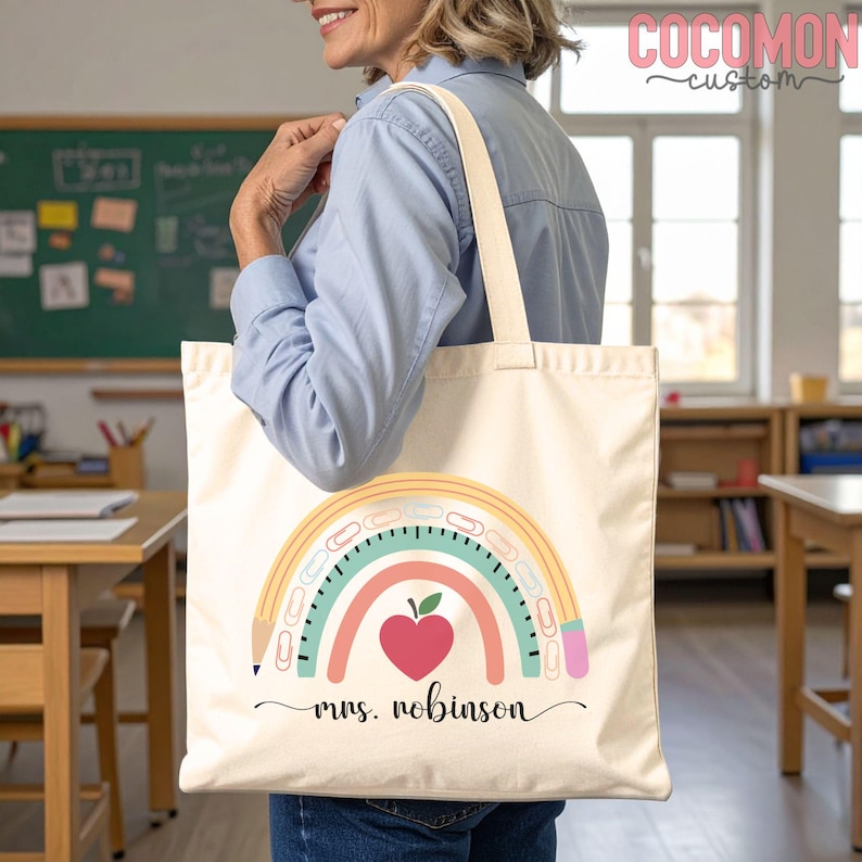 May include: Cream-colored tote bag with a rainbow pencil design and the text "mrs. robinson". The rainbow features a red apple heart, teal, pink, and yellow arcs. The bag has cream-colored handles and is being held by a person.