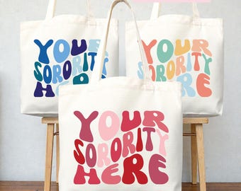 Op de afbeelding: Drie off-white canvas draagtassen met twee hengsels. Elke tas is voorzien van de tekst "YOUR SORORITY HERE" in een golvend retro-lettertype. De tekstkleuren variëren per tas, waaronder blauwtinten, pasteltinten en rood- en rozetinten.