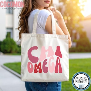 以下が含まれることがあります： ピンクと赤の色合いで「CHI OMEGA」の文字が入ったクリーム色のトートバッグ。バッグはシンプルなデザインで、2つのハンドルが付いており、丈夫な生地で作られています。バッグは人が持っています。