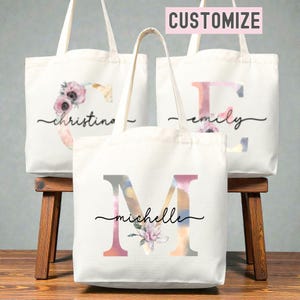 May include: Three white canvas tote bags with floral and initial designs. Each bag features a different initial and name, with the word "customize" in a pink box at the top. The bags are displayed on a wooden stool.