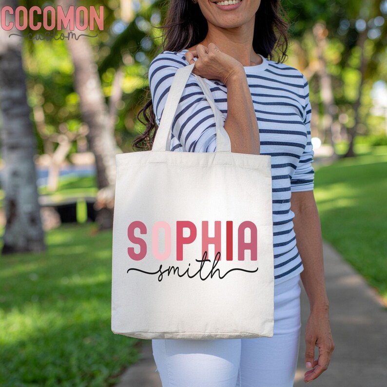 May include: A white canvas tote bag with the name "SOPHIA smith" printed in pink and red letters with a black cursive font. The bag is being held by a person wearing a striped shirt and white pants.