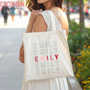 May include: Cream-colored tote bag with the name "EMILY" printed in a stacked design. The middle "EMILY" is in a pink and red color. The bag has a simple design and is carried over the shoulder.