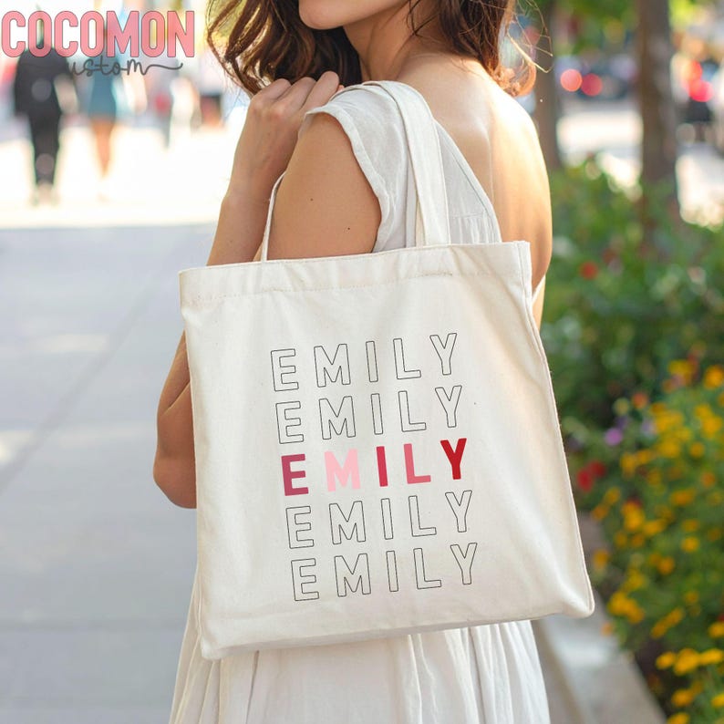 May include: Cream-colored tote bag with the name "EMILY" printed in a stacked design. The middle "EMILY" is in a pink and red color. The bag has a simple design and is carried over the shoulder.