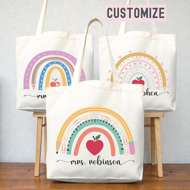 May include: Three white canvas tote bags with rainbow designs. Each bag features a colorful rainbow, a red apple, and a pencil. The bottom bag has the text "mrs. robinson" in a script font. The top right bag has the text "Customize" in a pink box.