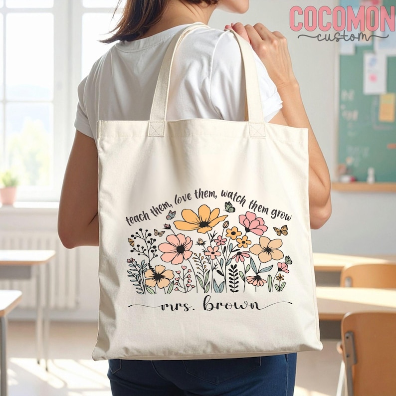 May include: Cream-colored tote bag with a floral design in pastel colors and the text "teach them, love them, watch them grow" and "mrs. brown". The bag has two handles and is being held over a person's shoulder.