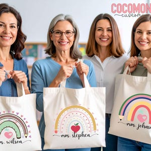 May include: Four women holding white tote bags with rainbow and apple designs. Each bag has a different name printed below the rainbow. The bags have a pencil design on the side of the rainbow and a heart in the center.