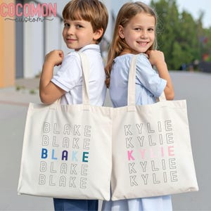 May include: Two off-white canvas tote bags with the names "Blake" and "Kylie" printed in a stacked design. The name "Blake" has blue and teal accents, while "Kylie" has pink accents. The bags have long handles.