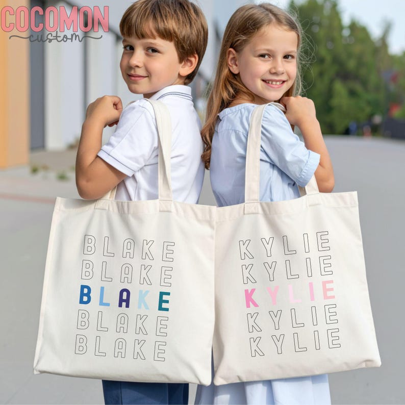 May include: Two off-white canvas tote bags with the names "Blake" and "Kylie" printed in a stacked design. The name "Blake" has blue and teal accents, while "Kylie" has pink accents. The bags have long handles.