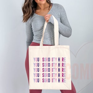 Op de afbeelding: Een witte canvas tote bag met de tekst "YOUR SORORITY HERE" die vijf keer wordt herhaald in verschillende kleuren, waaronder roze, paars en blauw.
