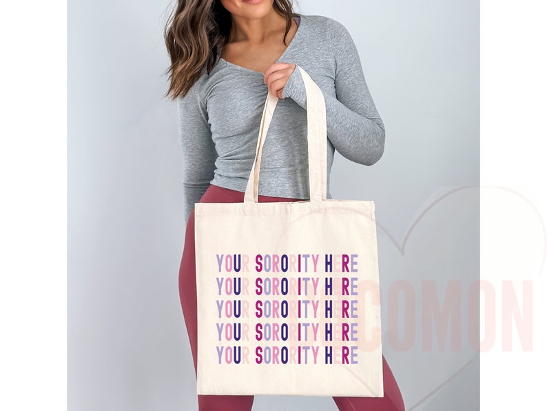 Op de afbeelding: Een witte canvas tote bag met de tekst "YOUR SORORITY HERE" die vijf keer wordt herhaald in verschillende kleuren, waaronder roze, paars en blauw.