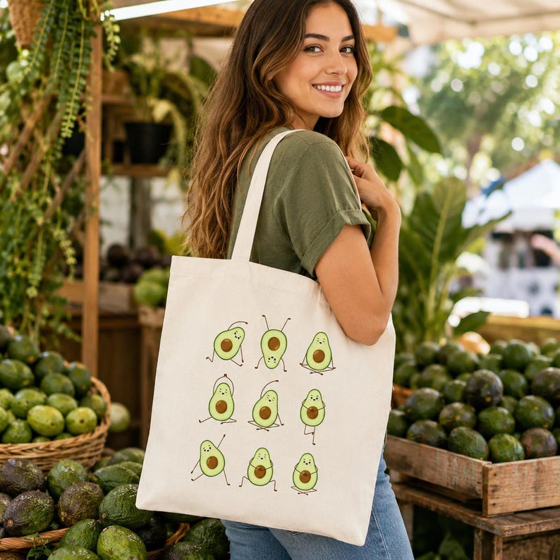 May include: Cream-colored tote bag featuring a repeating pattern of cartoon avocados in various yoga poses. The avocados are green with brown seeds and black outlines. The bag is held by a person in a green shirt.