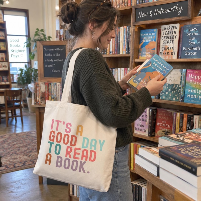May include: A white canvas tote bag featuring the multi-colored text "IT'S A GOOD DAY TO READ A BOOK." The bag has two shoulder straps and is constructed from a light-colored fabric, displaying a clear, bold, sans-serif font in various colors.