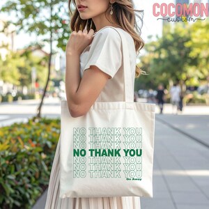 May include: Cream-colored tote bag with the text "NO THANK YOU" in a repeating pattern, with the words in green. The phrase "Go Away" is printed below the text. The bag is carried by a person.