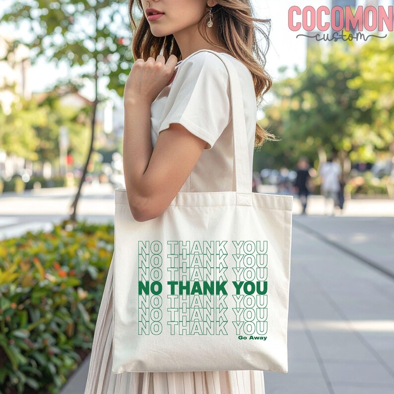 May include: Cream-colored tote bag with the text "NO THANK YOU" in a repeating pattern, with the words in green. The phrase "Go Away" is printed below the text. The bag is carried by a person.