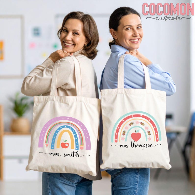 May include: Two canvas tote bags with rainbow designs, apples, and the names "Mrs. Smith" and "Mrs. Thompson". The bags have cream-colored fabric and feature colorful illustrations of rainbows, pencils, and stars. The bags are held by two people.