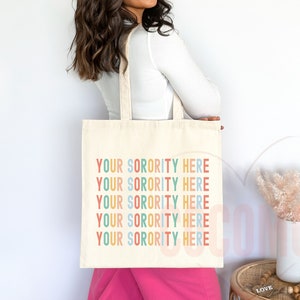 Op de afbeelding: Een witte tote bag met de tekst "YOUR SORORITY HERE" die vijf keer in regenboogkleuren wordt herhaald. De tekst is in een speels, casual lettertype.