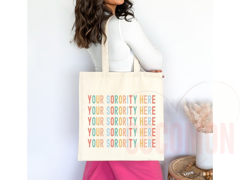Op de afbeelding: Een witte tote bag met de tekst "YOUR SORORITY HERE" die vijf keer in regenboogkleuren wordt herhaald. De tekst is in een speels, casual lettertype.