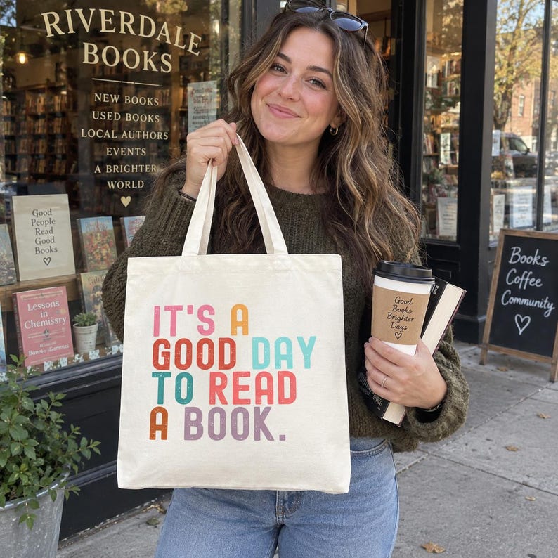 May include: A cream-colored canvas tote bag featuring the multi-colored text "IT'S A GOOD DAY TO READ A BOOK." The bag has two sturdy fabric handles and a flat rectangular shape, designed for carrying books and personal items with a clear, bold print.