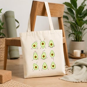 May include: A cream-colored tote bag featuring a playful design of avocados in yoga poses. The avocados are green with brown pits and are depicted in various yoga positions. The bag has long handles for carrying.