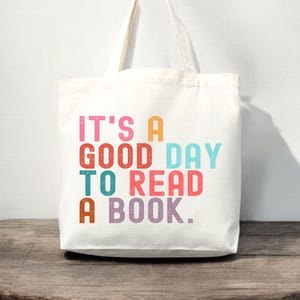 May include: A white canvas tote bag with two shoulder straps. The front features the multi-colored text IT'S A GOOD DAY TO READ A BOOK in a block font. The letters are printed in shades of pink, orange, red, light blue, teal, and purple.
