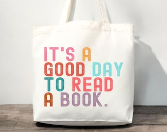 May include: A white canvas tote bag with two shoulder straps. The front features the multi-coloured text IT'S A GOOD DAY TO READ A BOOK in a block font. The letters are printed in shades of pink, orange, red, light blue, teal, and purple.