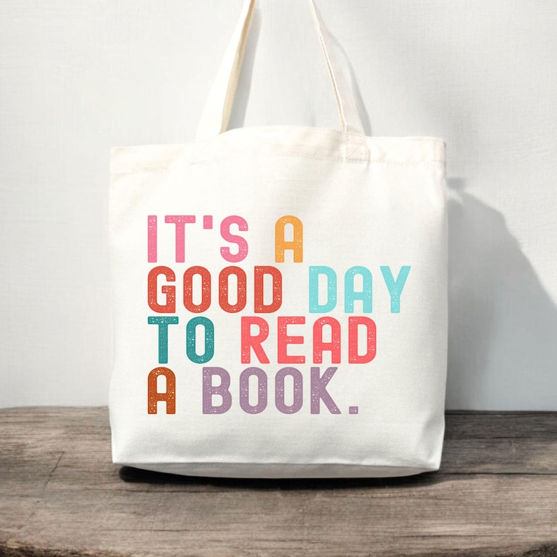 May include: A white canvas tote bag with two shoulder straps. The front features the multi-colored text IT'S A GOOD DAY TO READ A BOOK in a block font. The letters are printed in shades of pink, orange, red, light blue, teal, and purple.