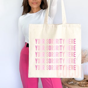 Op de afbeelding: Een witte canvas tote bag met de tekst "YOUR SORORITY HERE" vijf keer herhaald in roze letters, met een verloop van lichtroze naar donkerroze.