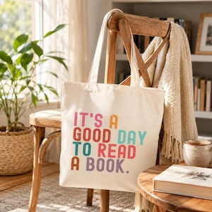 May include: A cream-colored canvas tote bag featuring the multi-colored text "IT'S A GOOD DAY TO READ A BOOK." The bag has two shoulder straps and a rectangular shape, with the printed lettering displayed in shades of pink, orange, teal, and purple.