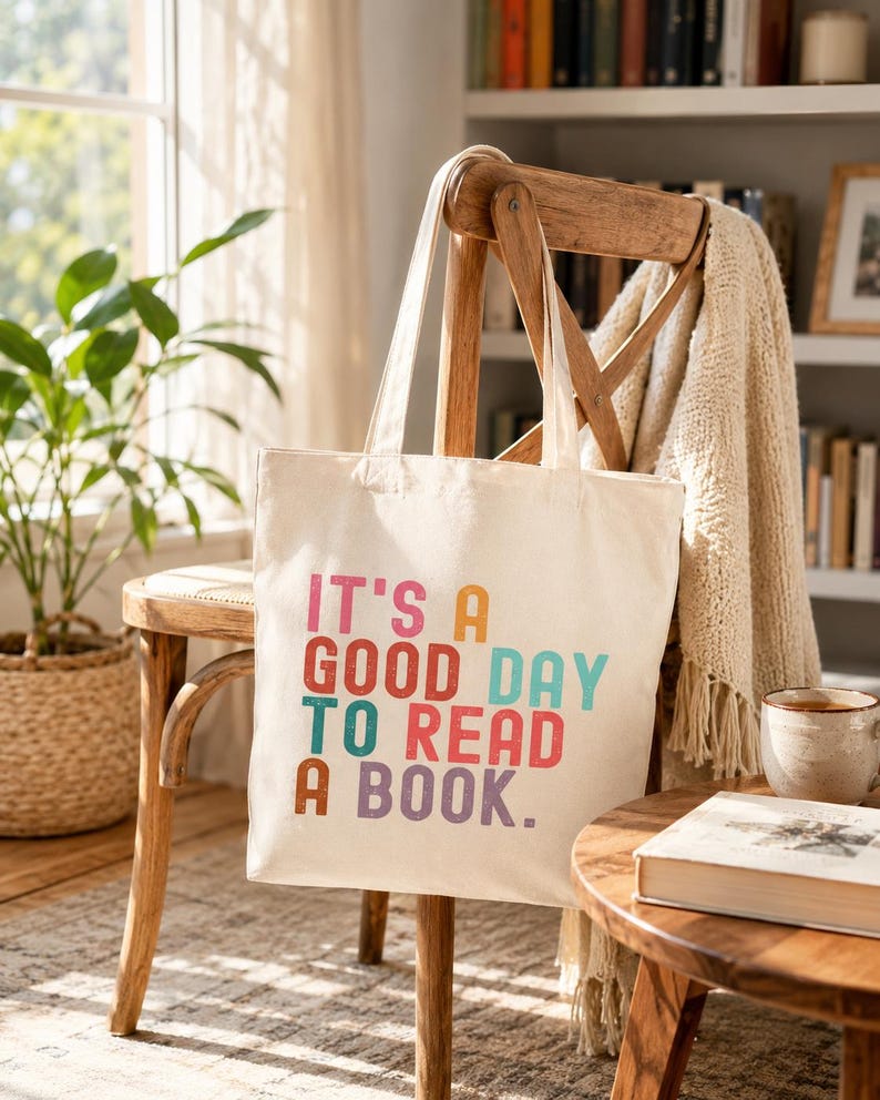 May include: A cream-colored canvas tote bag featuring the multi-colored text "IT'S A GOOD DAY TO READ A BOOK." The bag has two shoulder straps and a rectangular shape, with the printed lettering displayed in shades of pink, orange, teal, and purple.