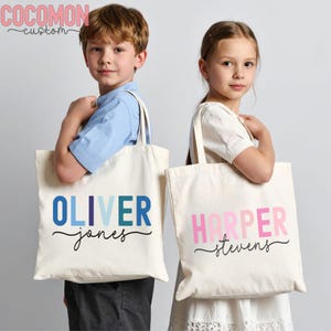May include: Two cream-colored tote bags with personalized names. One bag has "OLIVER" in blue hues and "jones" in black script. The other bag has "HARPER" in pink hues and "stevens" in black script. The bags have long handles.