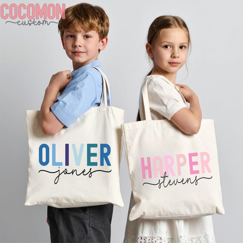 May include: Two cream-colored tote bags with personalized names. One bag has "OLIVER" in blue hues and "jones" in black script. The other bag has "HARPER" in pink hues and "stevens" in black script. The bags have long handles.