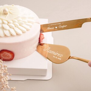 A hand is using a gold cake server to cut a white cake decorated with a strawberry on top.