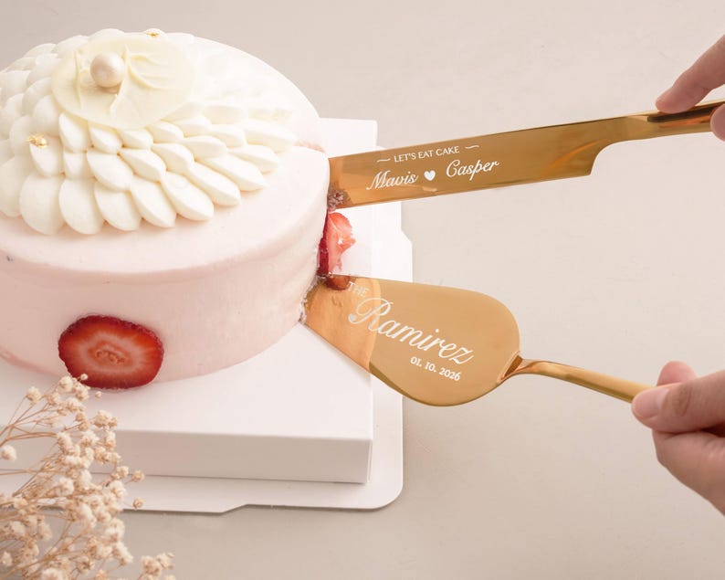 A hand is using a gold cake server to cut a white cake decorated with a strawberry on top.