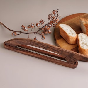 May include: A dark brown wooden bread slicer guide with a serrated edge, engraved with the text Home Is Where Bread Is Warm and Baked with Love, Just for Victoria, featuring the initials S L and a small wheat stalk graphic on the side.