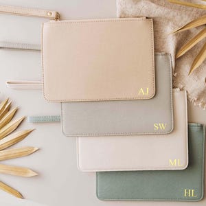 May include: Four zippered pouches in different colors and materials. The pouches are beige, gray, white, and green. Each pouch has a gold-colored zipper and a gold-colored monogram on the front. The monograms are AJ, SW, ML, and HL.