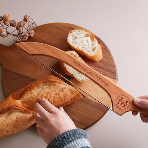 May include: A wooden bread slicer with a thin metal wire, featuring the engraved text "Made for Cozy Mornings" and a circular monogram with the letter M and the name "MICHELLE" inside, positioned over a loaf of bread on a round wooden board.