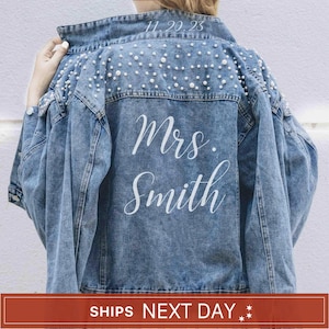 May include: A blue denim jacket featuring white pearl embellishments across the upper back and shoulders. The text 11.20.23 is printed near the collar, while the words Mrs. Smith are printed in a white script font across the center back panel.