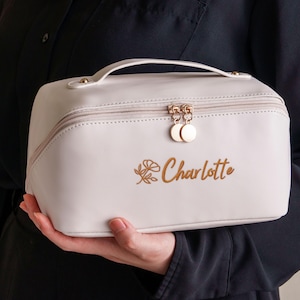 Puede incluir: Un neceser rectangular rosa claro con asa y cierre de cremallera. La bolsa tiene herrajes dorados y el nombre "Charlotte" está impreso en oro con un diseño floral.