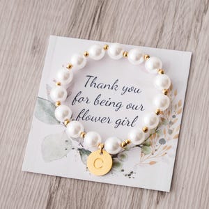 May include: A white pearl bracelet with gold accents and a gold charm with the letter "C" rests on a card. The card reads "Thank you for being our flower girl" with floral accents. The bracelet is a charming gift.