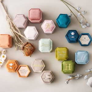 May include: A collection of hexagonal velvet ring boxes in various colors including pink, blue, green, and orange. Some boxes are closed, while others are open, revealing rings or personalized text printed in gold or white on the lids and inserts.