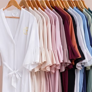 May include: A collection of elegant, soft-looking robes in a variety of colors, hanging on wooden hangers. The robes have a wrap-around design with wide sleeves and a tie belt. The first robe is white with gold embroidery.