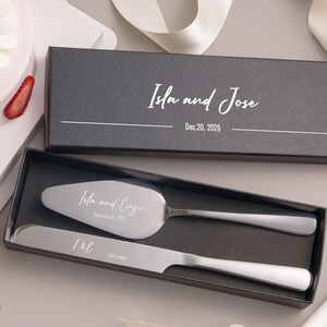 A black box containing a silver knife and a silver cake server, with a white ribbon and a strawberry decoration.