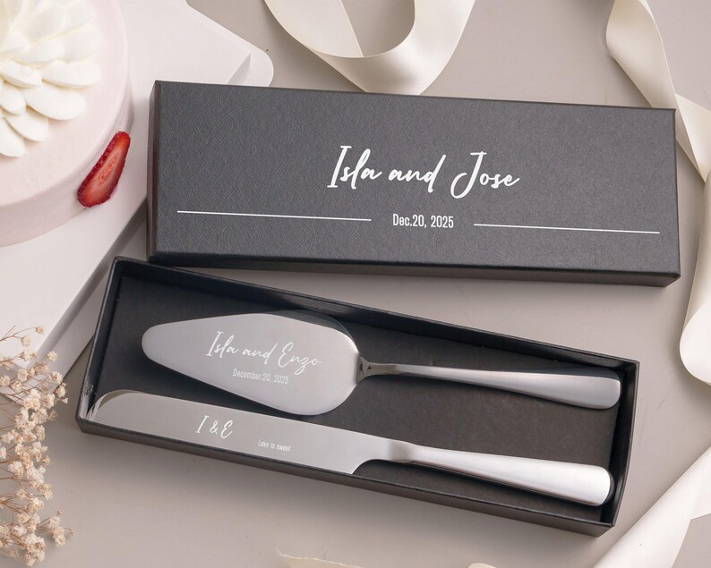 A black box containing a silver knife and a silver cake server, with a white ribbon and a strawberry decoration.